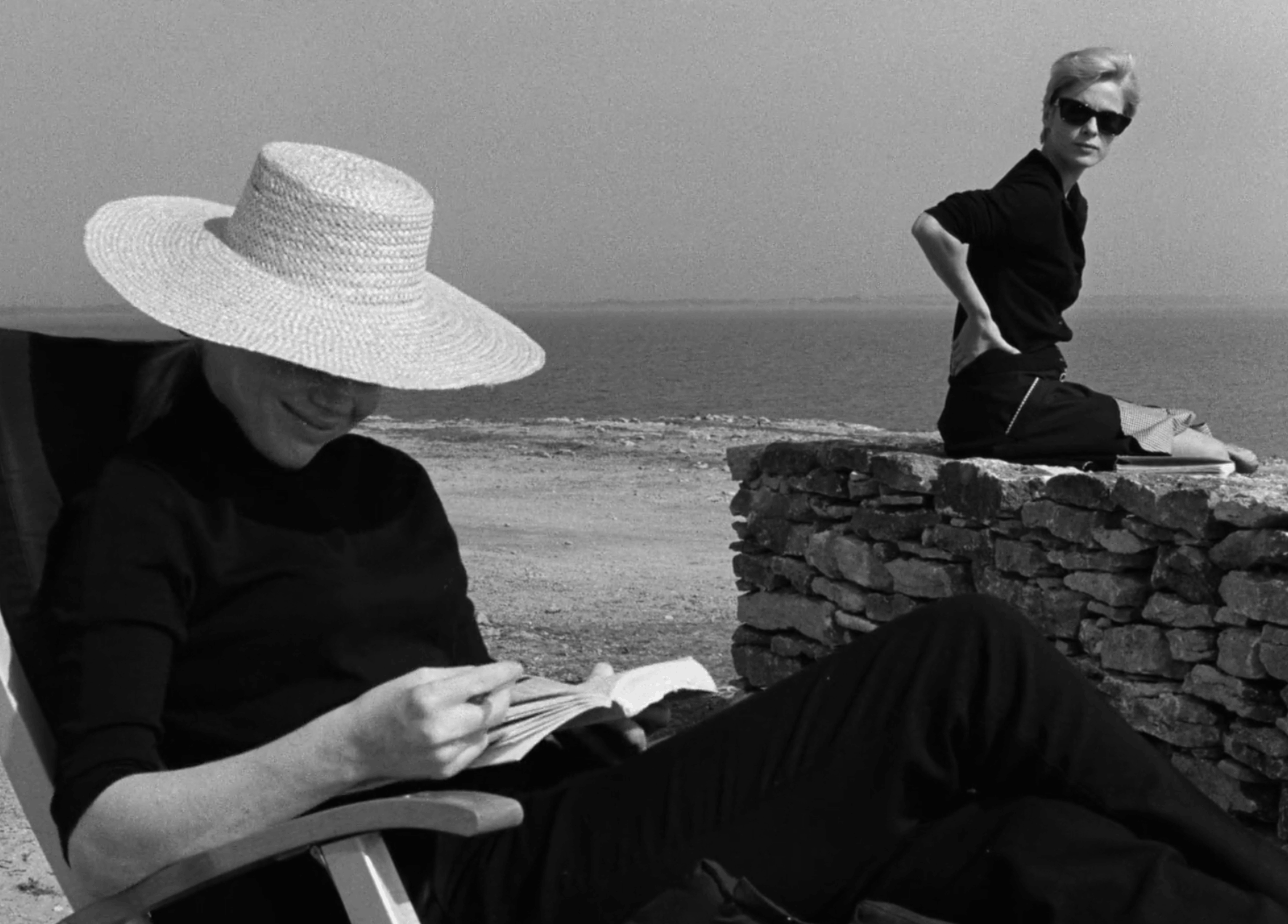Alma legge un libro mentre Elisabeth osserva in silenzio, sulla spiaggia in Persona di Ingmar Bergman.